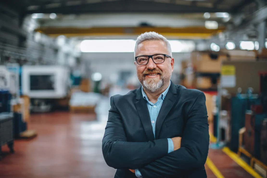 A masculine person with short grey hair and beard, wearing a suit, standing with arms folded smiling at the camera, in front of a slightly blurred workshop