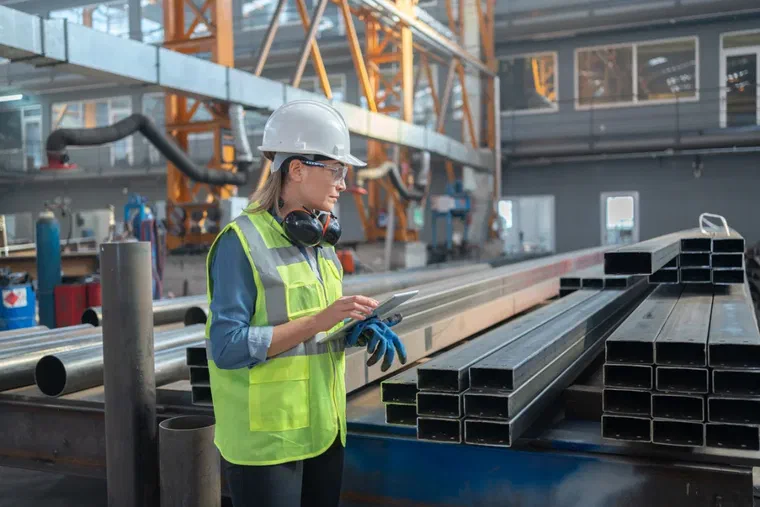 A stock controller in high-visibility workwear checking steel tube and pipe inventory on a tablet in a metals warehouse