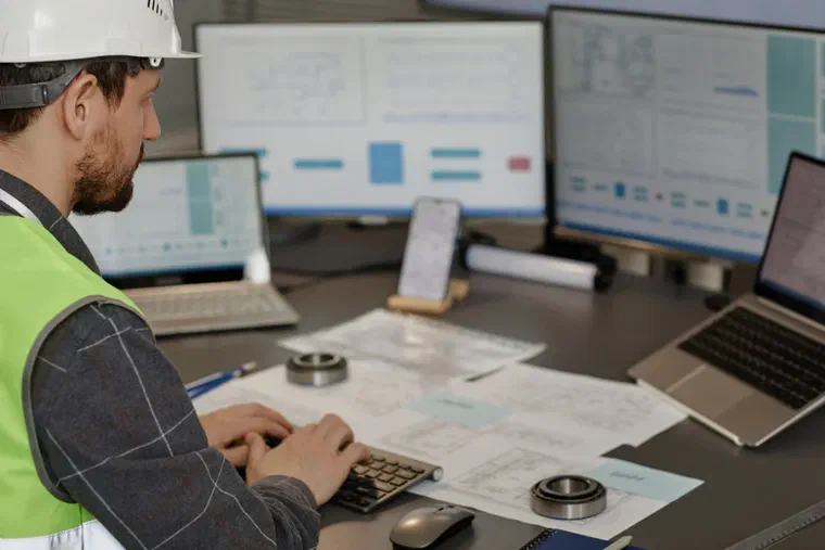An operations planner in a hard hat reviewing production dashboards across multiple screens at a manufacturing facility